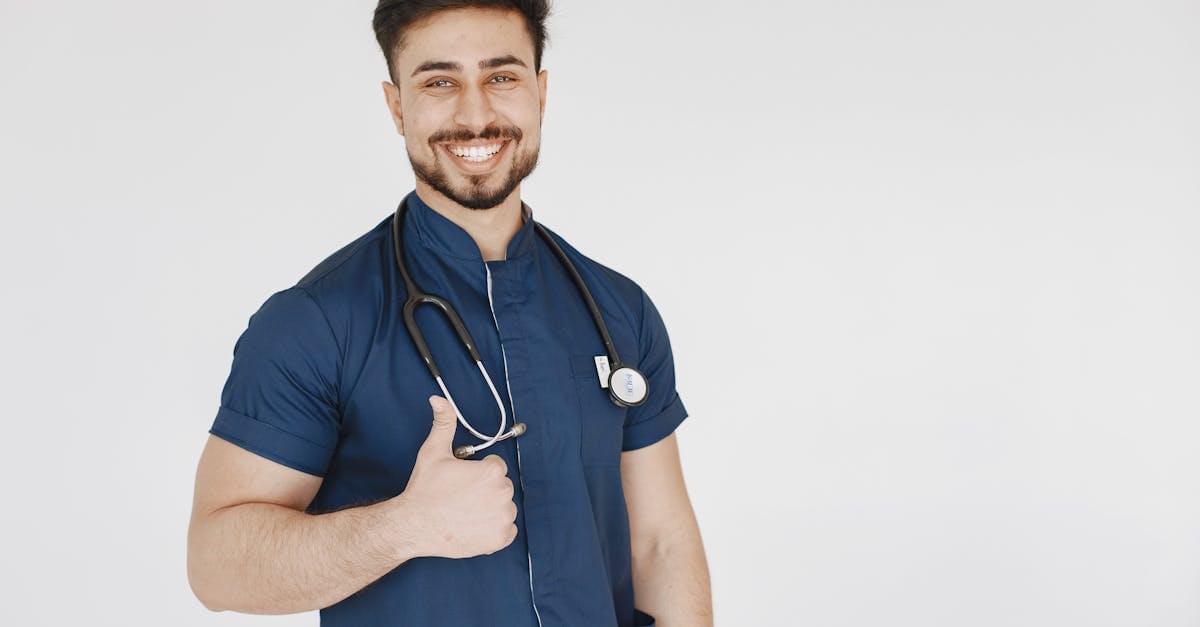 Confident male doctor in blue scrubs with a stethoscope smiling and giving a thumbs up.